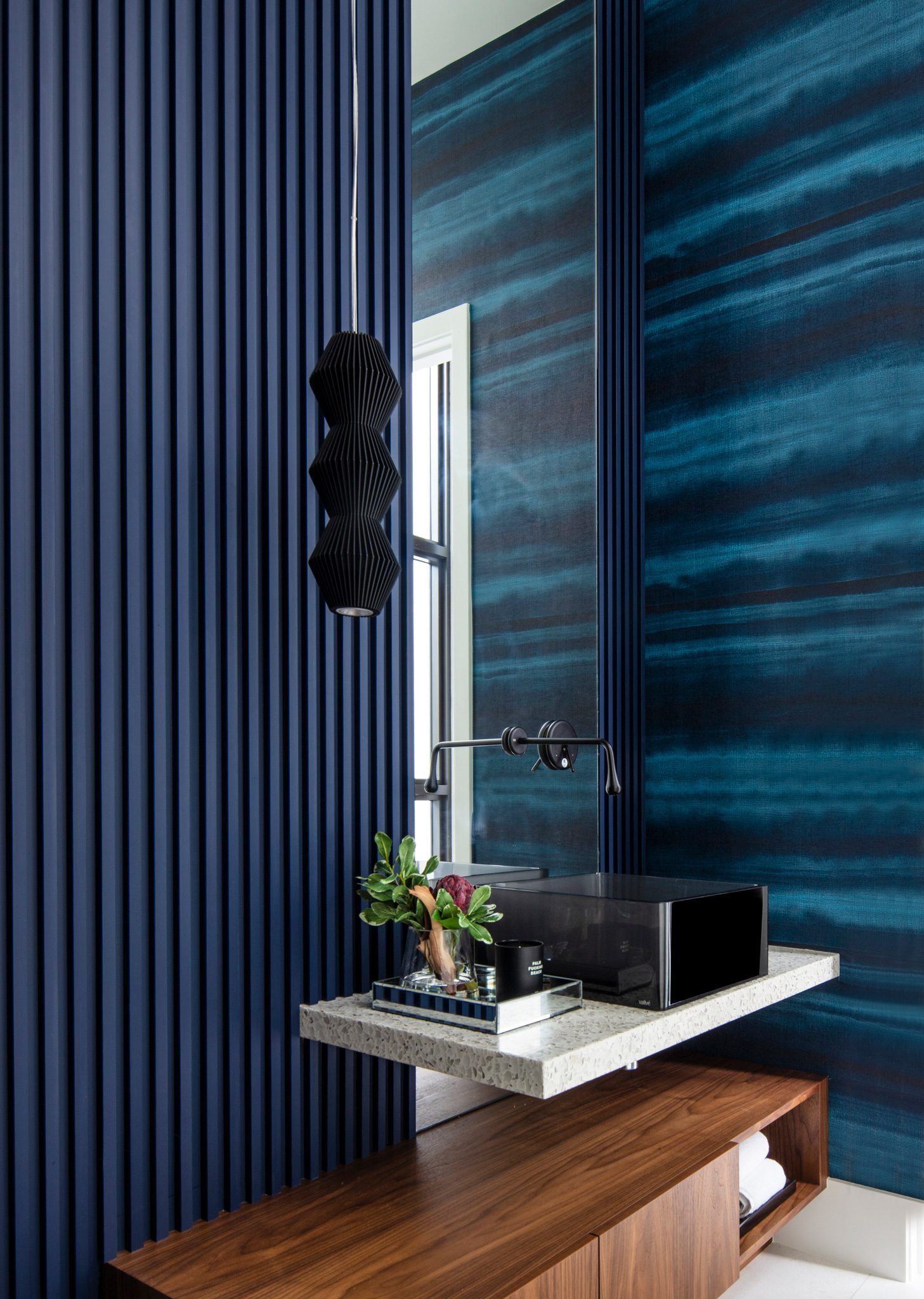 Modern bathroom with dark blue walls, a floating sink with a rectangular mirror above, a black pendant light, and a wooden vanity below. A small tray with decorative items sits on the sink counter.