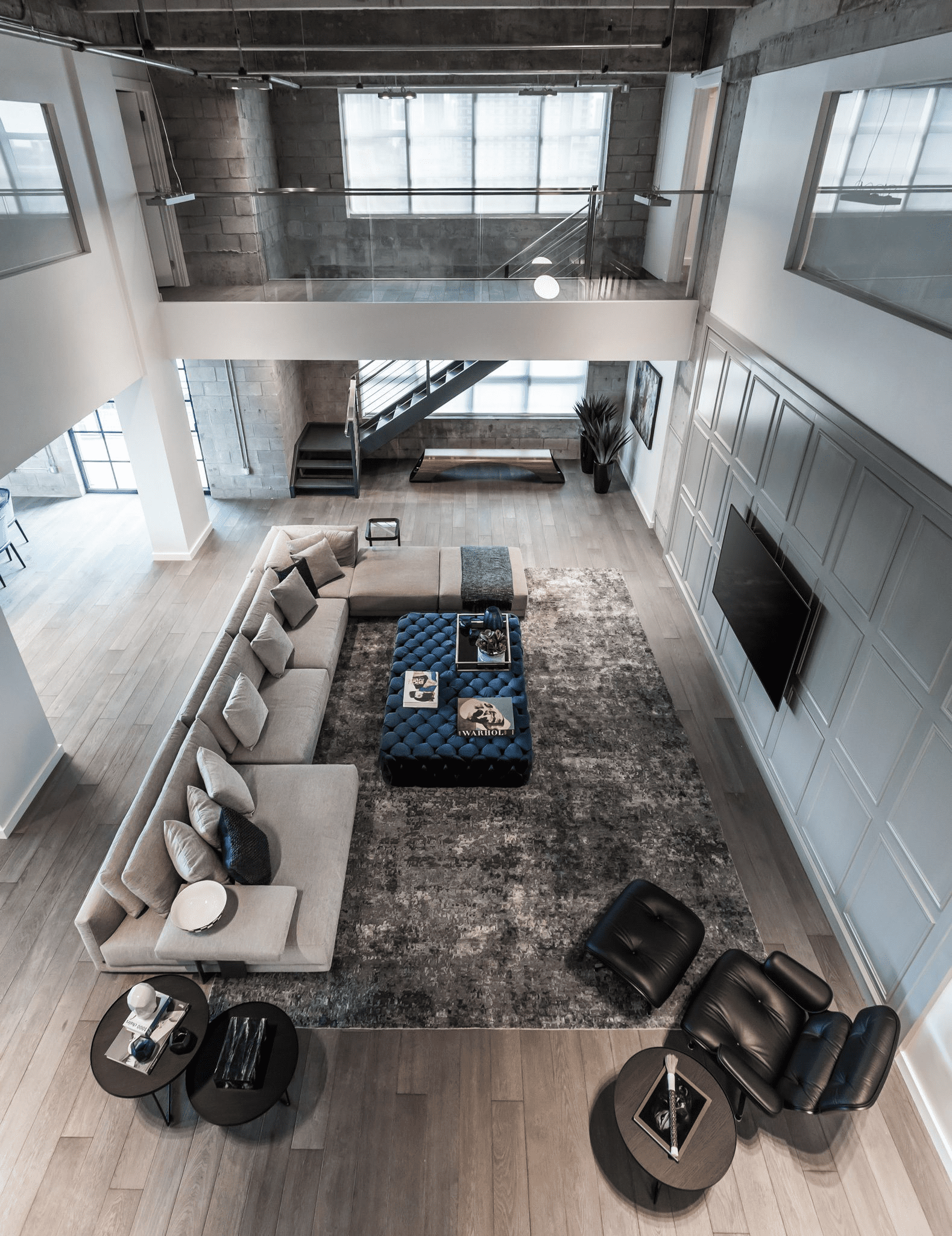 A modern, open-concept loft with high ceilings and large windows. It features a sectional sofa, a black recliner, and a coffee table on a gray rug in the living area. Stairs lead to an upper level.