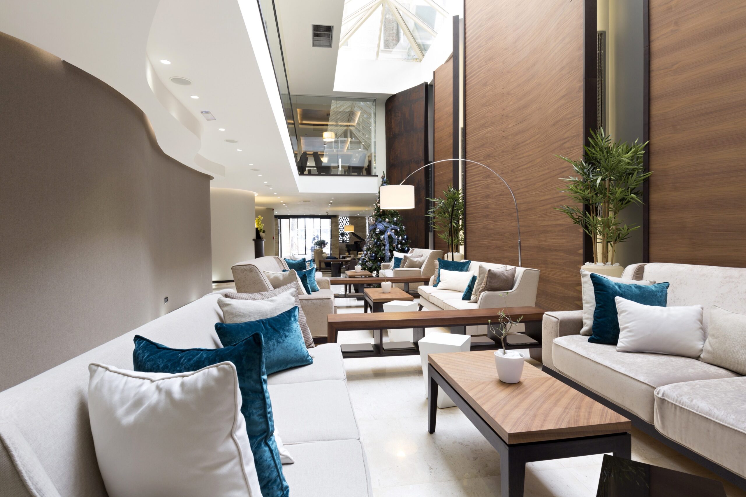 A modern hotel lounge with white sofas, teal cushions, wooden tables, plants, and a high ceiling.